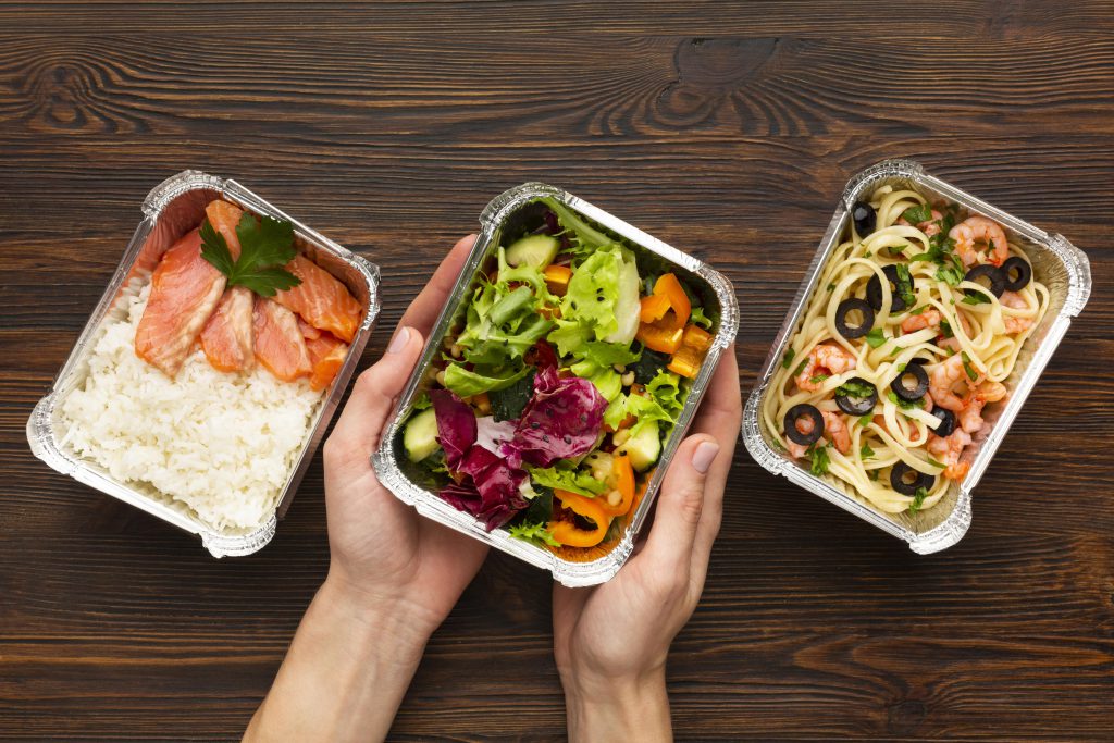 Propozycje posiłków z diety pudełkowej bez glutenu i laktozy fotografowane z lotu ptaka. Dłonie trzymające jeden pojemnik z posiłkiem.