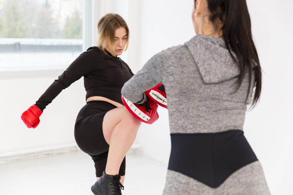 Kurs samoobrony dla kobiet - kobieta wykonująca kopnięcie podczas treningu boksu, trzymająca rękawice, a druga kobieta przygotowuje się do przyjęcia ciosu.