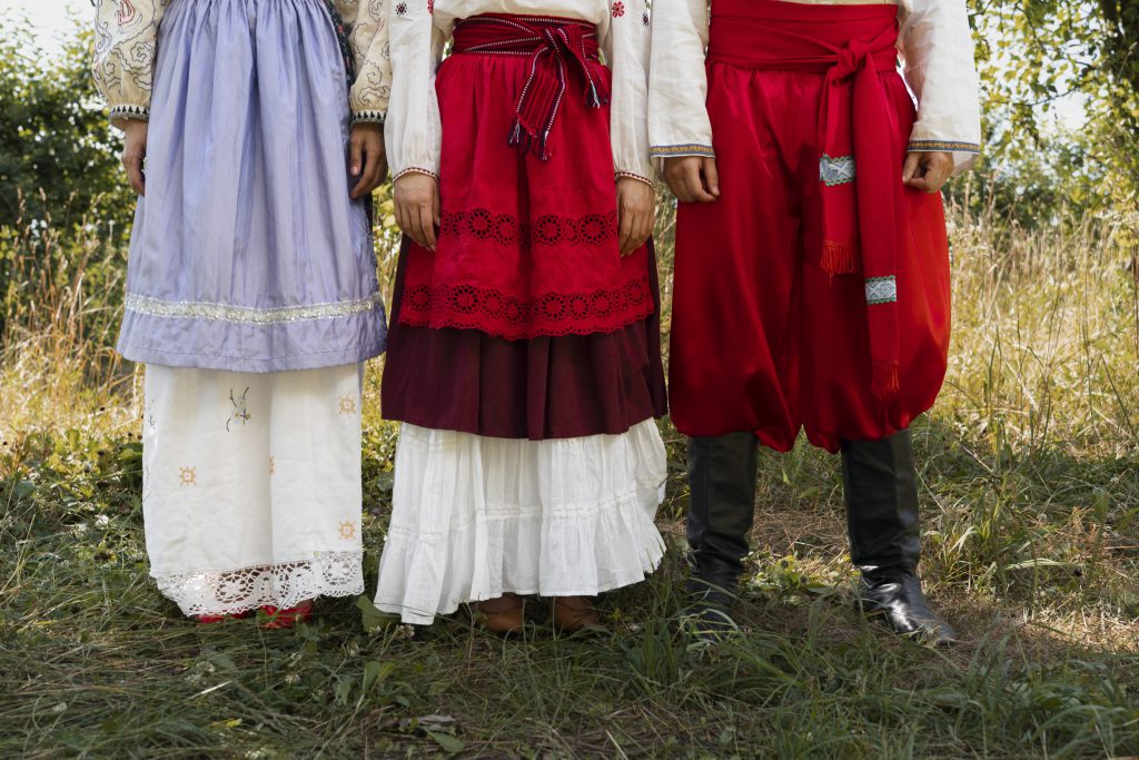 Trzy osoby w tradycyjnych strojach folkowych, każda w kolorowej spódnicy folkowej, tańczące w plenerze