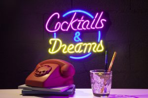 Kolorowy neon w biurze z napisem „Cocktails & Dreams”, świecący na ciemnej ścianie obok retro telefonu i szklanki z lodem
