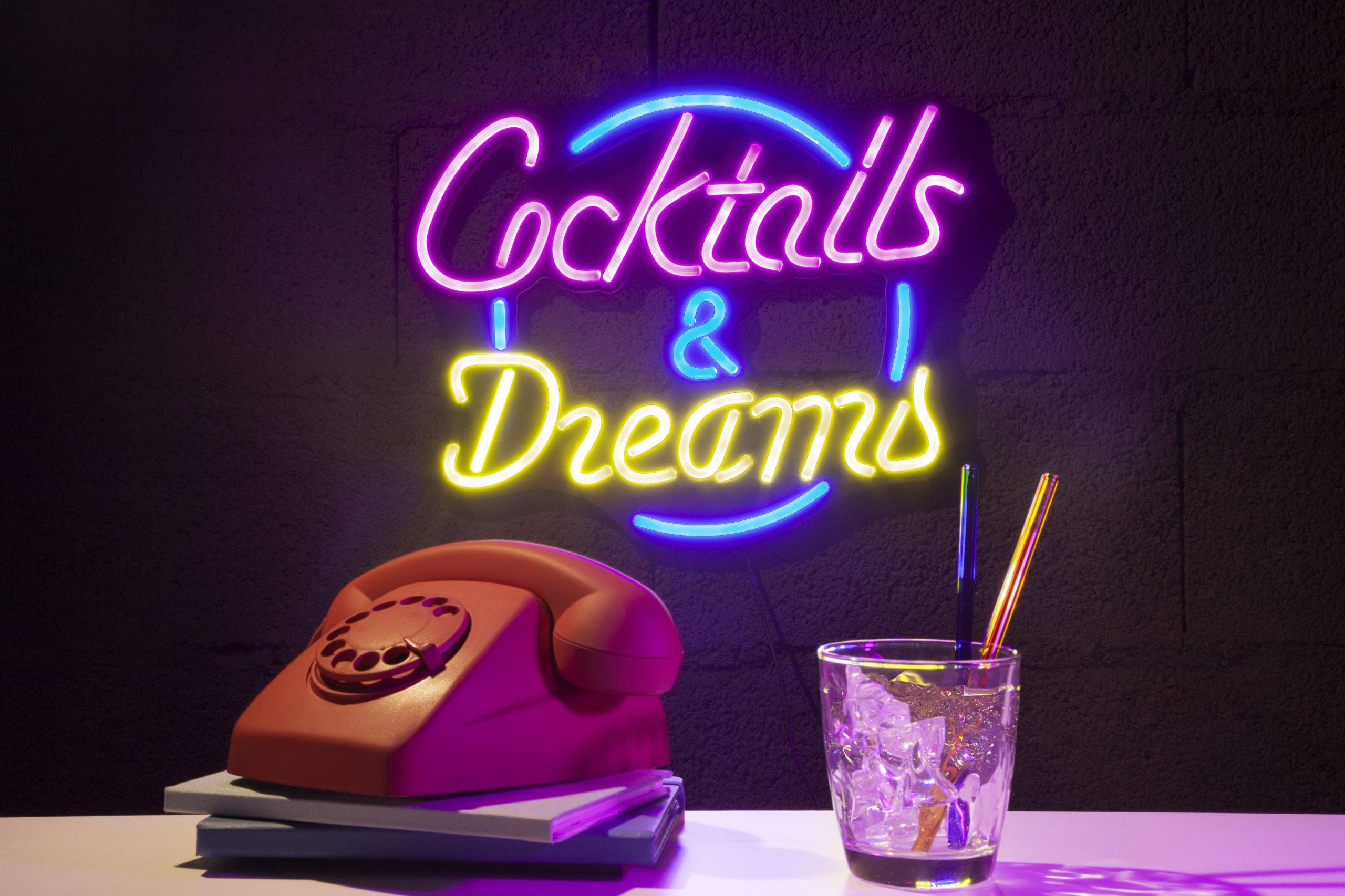 Kolorowy neon w biurze z napisem „Cocktails & Dreams”, świecący na ciemnej ścianie obok retro telefonu i szklanki z lodem