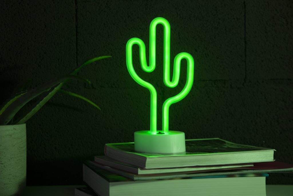 Zielony neon w biurze przedstawiający kaktus, ustawiony na stosie książek obok doniczki z rośliną