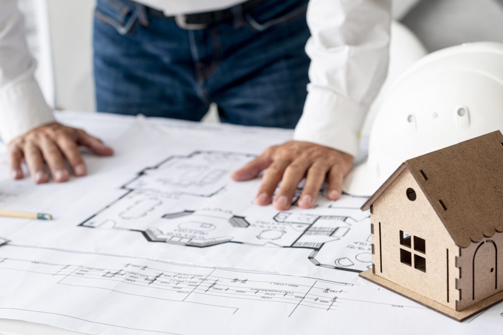 Projekt domu inwestycyjnego na stole, z planszą architektoniczną, makietą domu oraz kaskiem budowlanym, reprezentujący inwestycje w nieruchomości