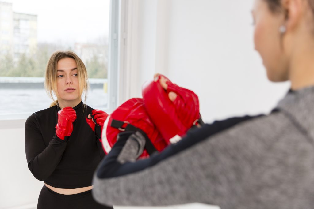Kurs samoobrony dla kobiet - kobieta przygotowująca się do treningu boksu, trzymająca rękawice w czerwonym kolorze, patrzy na trenującą partnerkę.